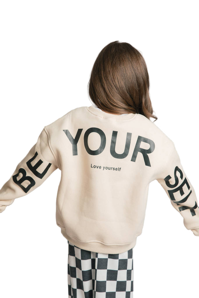 Person wearing a beige sweatshirt with text on the back and sleeves, standing against a light gray background.