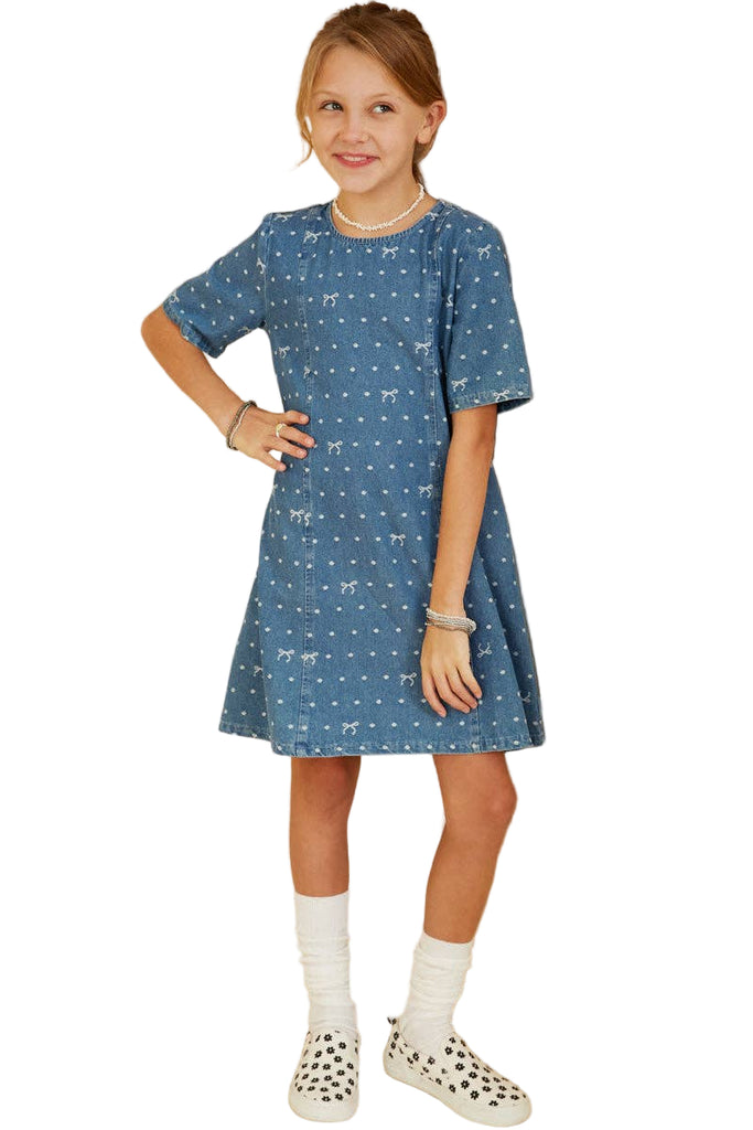 Young girl wearing a blue dress with a pattern, standing in front of a wall with framed pictures.